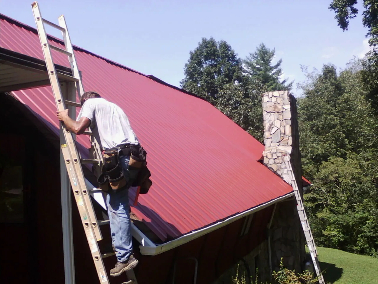 Roofer on a ladder beside a red metal roof and stone chimney — Soffit & Fascia Repair by Roofing Brothers in Coleman